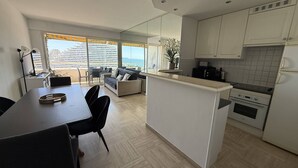 Une cuisine moderne avec des meubles blancs, un coin repas comprenant une table et des chaises, et une vue sur la mer grâce à de grandes fenêtres.