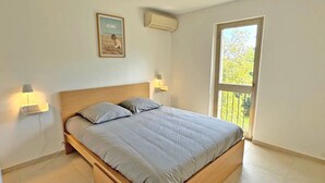 A bedroom with a bed, two bedside lamps, a wall-mounted air conditioner, a window with a view of trees, and a framed picture on the wall. - Apartment for 6people - Air conditioning - Litlle sea view - Wifi - Sainte Maxime (STE MAXIME)