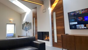 A modern living room with a fireplace, a black leather sofa, a wooden cabinet, and a mounted TV displaying sports channels.