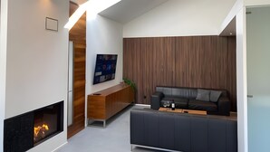 A modern living room with a fireplace, a wooden ceiling beam, a dark sofa, and a wooden slatted wall.