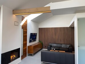 Una sala moderna con chimenea, una viga de madera en el techo, un sofá oscuro y un panel de listones de madera.