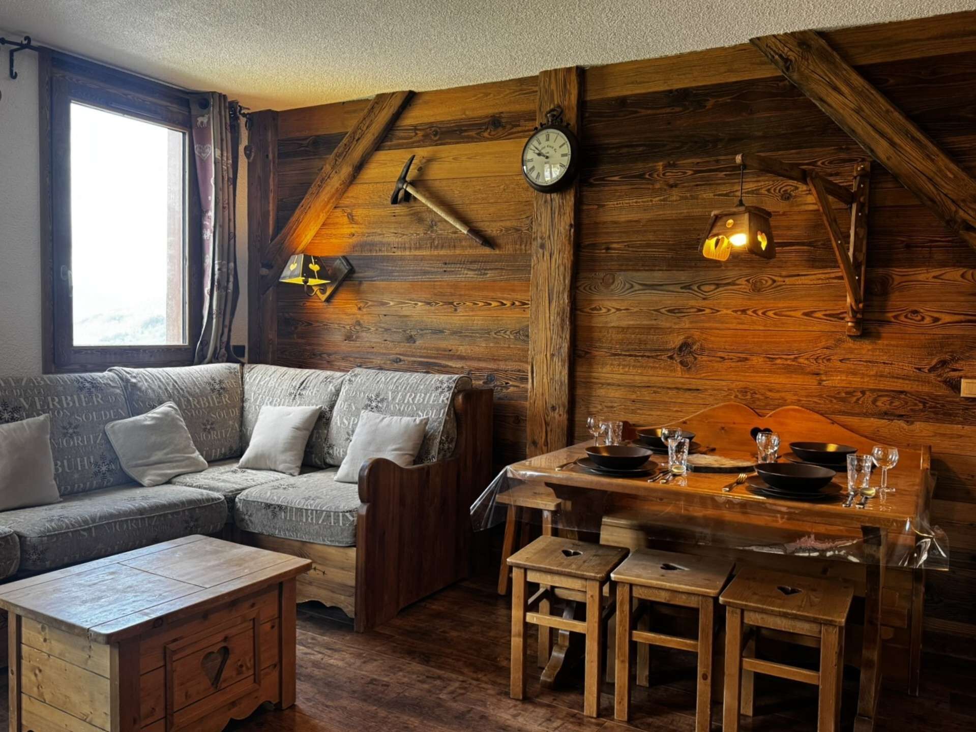 A rustic wooden interior with a sofa, dining table, and wooden stools. A clock and wall-mounted lamp are visible.