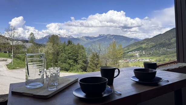 Une table sur laquelle se trouvent un verre d’eau, deux tasses noires et un verre, le tout posé sur une surface en bois, avec en toile de fond un paysage montagneux sous un ciel dégagé.