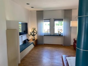 Une chambre moderne avec un plancher en bois, un téléviseur à écran plat fixé au mur, une petite table ornée d’un vase de fleurs et une fenêtre avec des rideaux.