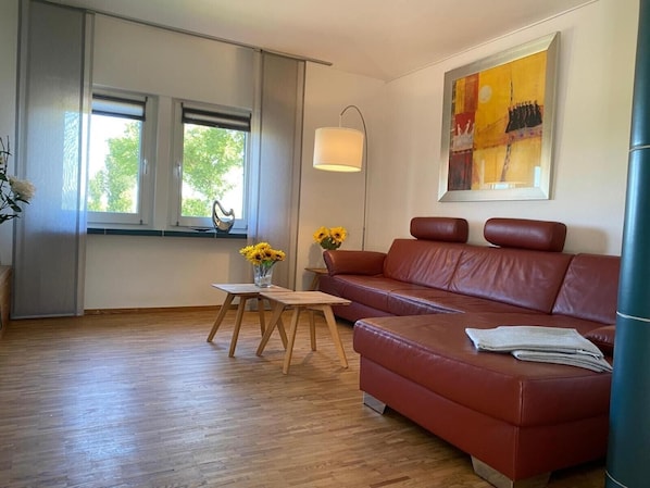 Un salon moderne avec un canapé en cuir, une petite table en bois ornée d’un vase de tournesols et une grande fenêtre donnant sur un paysage arboré.