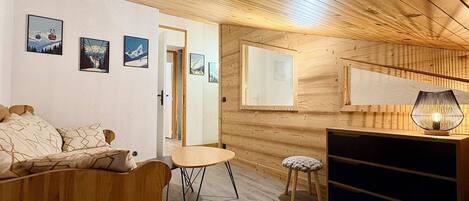 A cozy room with a wooden ceiling, a bed, a small table, a stool, and a TV stand.