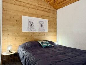Ett sovrum med ett träpanelat tak, en säng med ett mörkt täcke, ett litet runt bord med en lampa och en vägg med två djurmotiv.