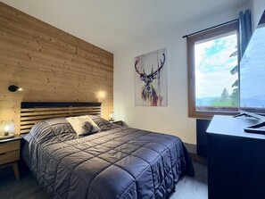 A bedroom with a wooden slat headboard, a bed with a quilted cover, a nightstand with a lamp, a painting of a deer, and a window with a view of trees. - Cheval Noir 32 - Charming 3-room apartment (LES AVANCHERS VALMOREL)