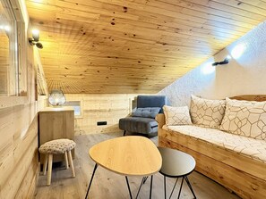 A cozy attic room with a wooden ceiling, a sofa, a small table, and a chair. - Cheval Noir 32 - Charming 3-room apartment (LES AVANCHERS VALMOREL)