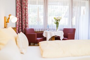 Habitación de hotel con una cama, dos sillas rojas, una mesita con un florero y una ventana con cortinas traslúcidas.