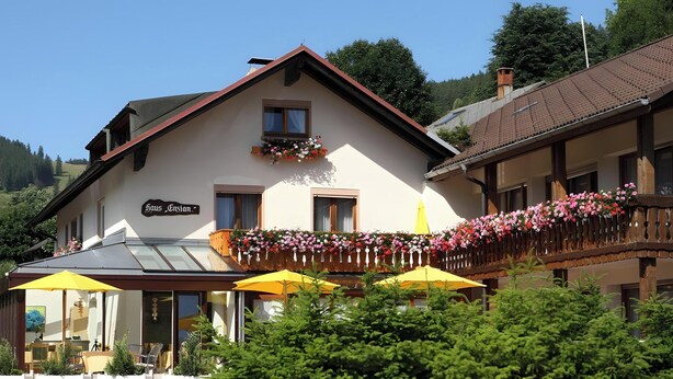 A building with a sign reading 'Haus Gvion,' a balcony with flowers, and outdoor seating with umbrellas. - Hotel-Pension Enzian – Double Room with Balcony or Terrace, Breakfast Included (Todtnau)