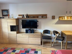 A modern living room with a flat-screen TV, wooden furniture, a bookshelf, and a dining area. - Studio 'Inzell mit Mountain View' with Balcony and Wi-Fi (Inzell)