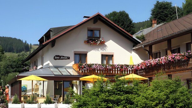 A building with a sign reading 'Haus Gvion,' a balcony with flowers, and outdoor seating with umbrellas.