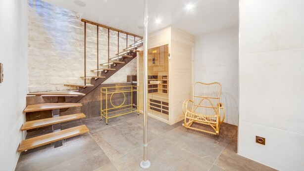 Un interior moderno con una escalera de madera, una sauna con paredes de vidrio y una silla de mimbre.
