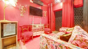 Una habitación con una decoración en tonos rosados, un televisor, una cama, un aparador y dos sillas.
