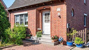 A red brick house with a white door, a small porch, and potted plants. - Apartment 'Gemütliche' with Shared Terrace, Shared Garden and Wi-Fi (Cuxhaven)