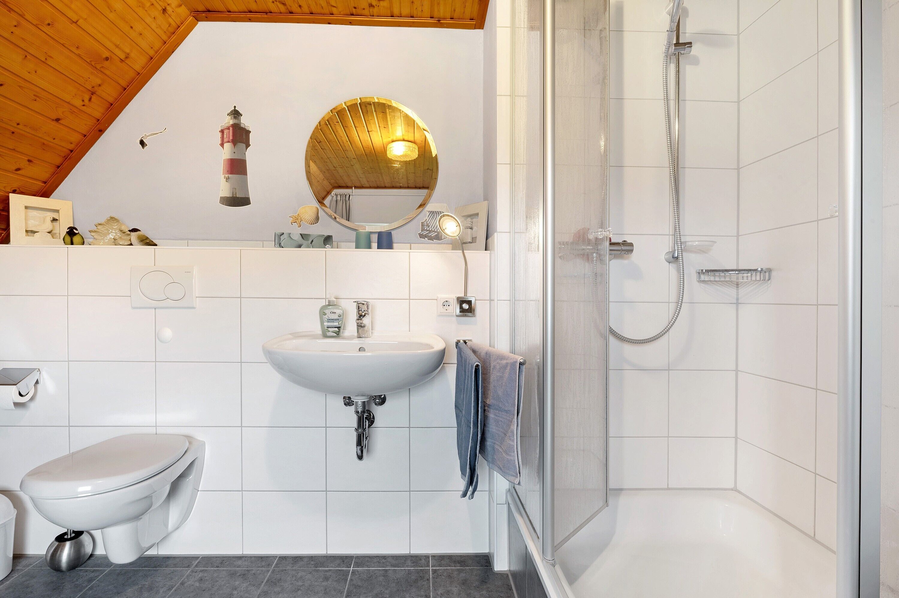 Ein Badezimmer mit weißem Waschbecken, Toilette und Dusche, Holzboden und einem runden Spiegel.
