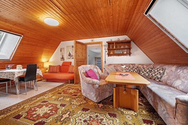 A cozy room with a wooden ceiling, a dining table with chairs, a sofa, and a patterned rug.