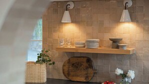 A kitchen with a tiled backsplash, two pendant lights, a wooden shelf with dishes, a rustic cutting board, and a small vase with flowers. - Woods Cove Inn (Laguna Beach)