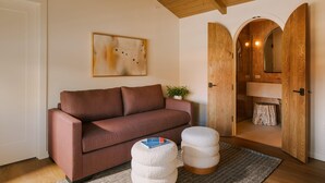 A room with a brown sofa, two round ottomans, a rug, a painting, and a wooden door leading to a bathroom. - Woods Cove Inn (Laguna Beach)