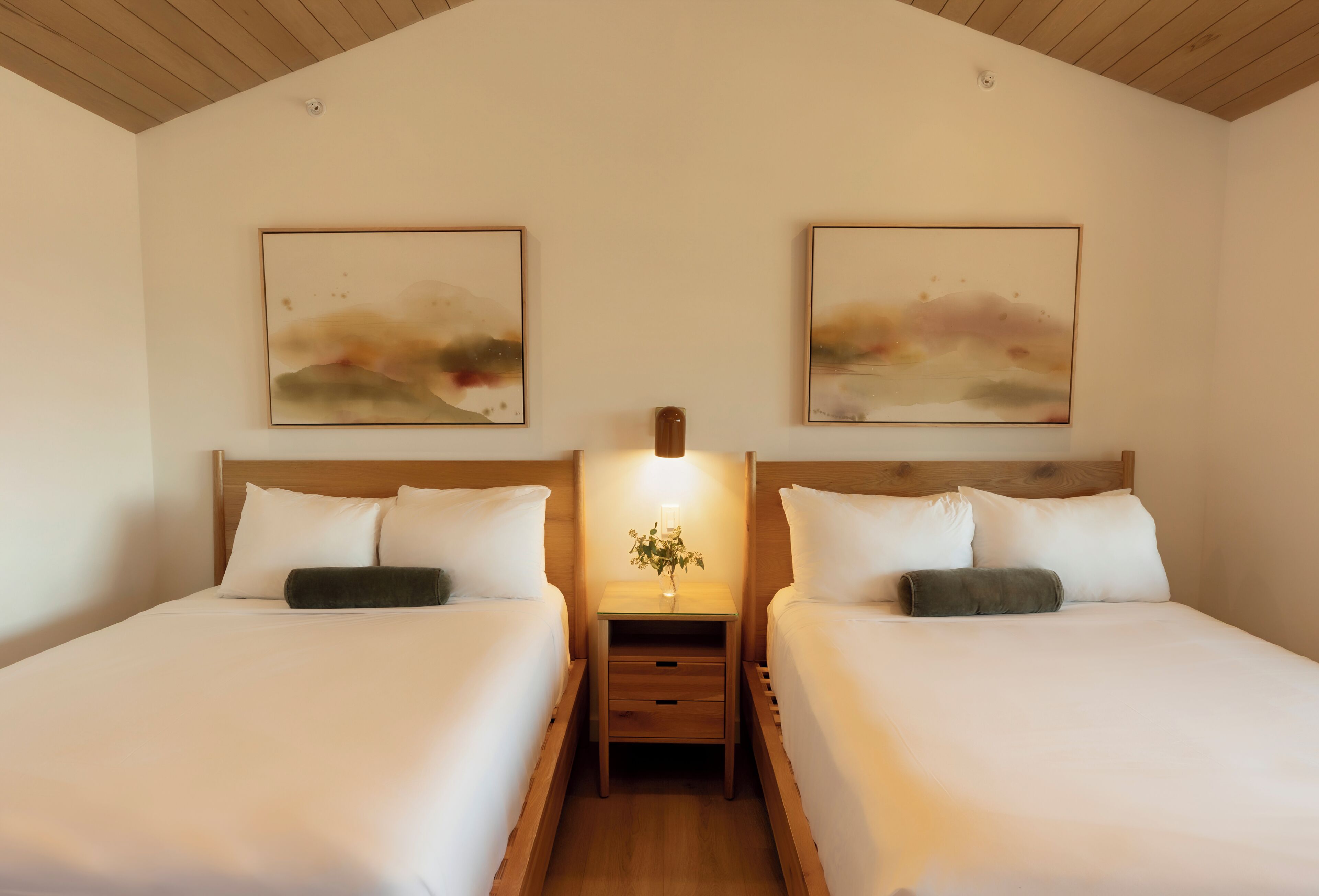 Une chambre d’hôtel avec deux lits, une table de chevet avec une lampe et des tableaux abstraits accrochés au mur.