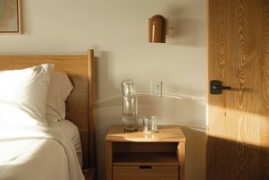Una mesita de noche de madera con un jarro y un vaso de vidrio, una puerta de madera y una cama con ropa blanca.