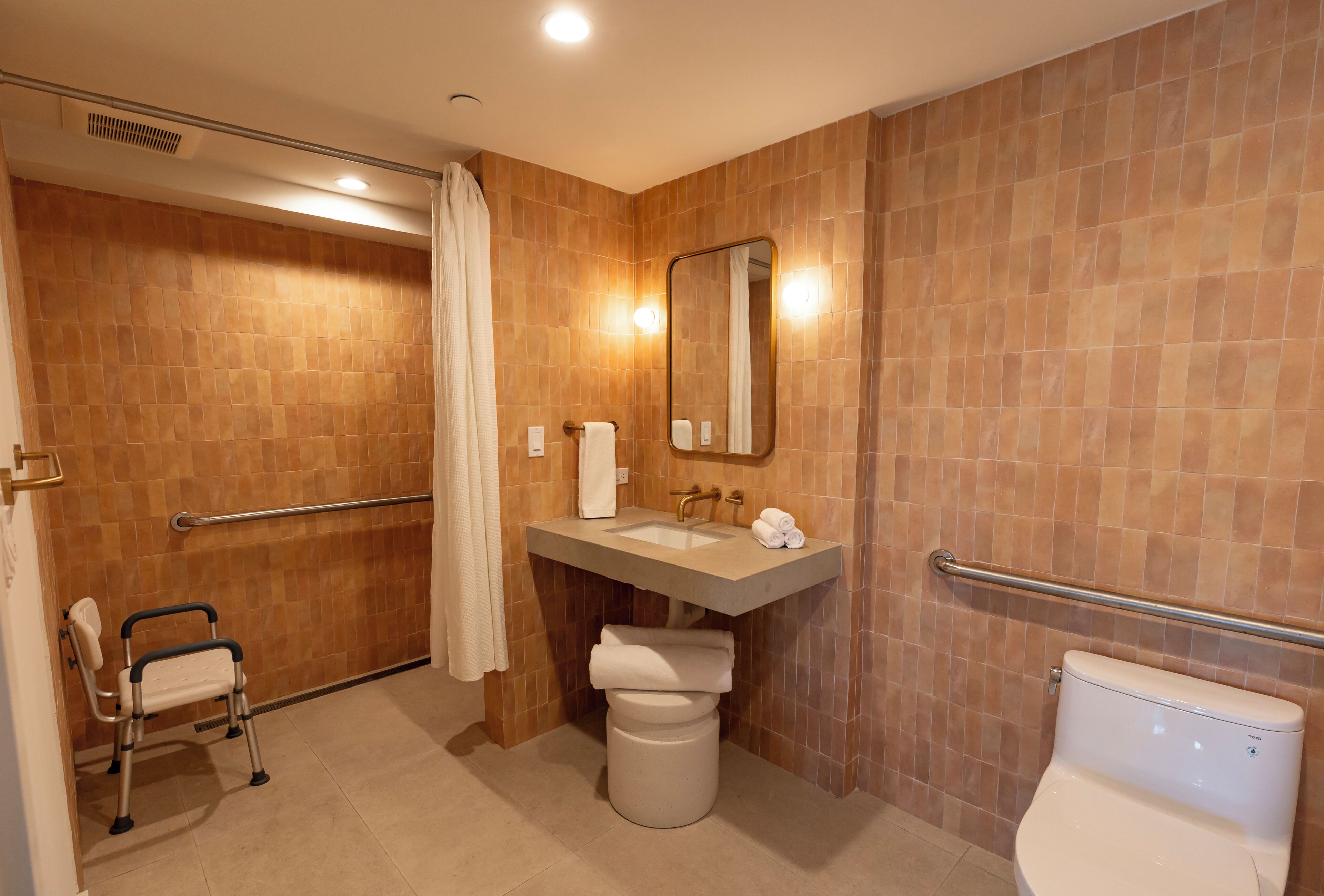 Une salle de bain équipée d’une douche, de toilettes, d’un lavabo avec un miroir et d’un banc accessible aux personnes en fauteuil roulant.