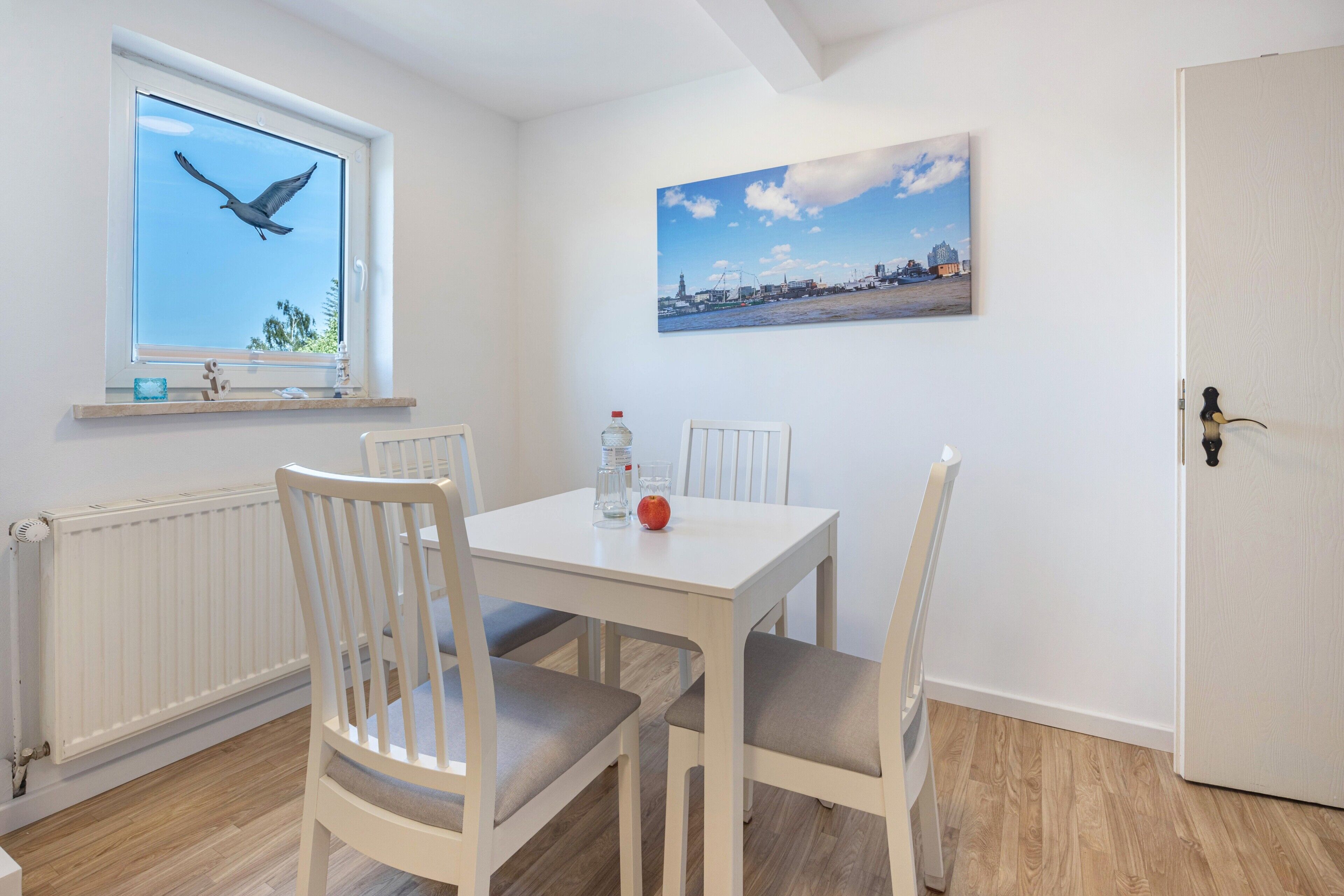 A dining area with a white table and chairs, a window with a bird, a painting, and a door.
