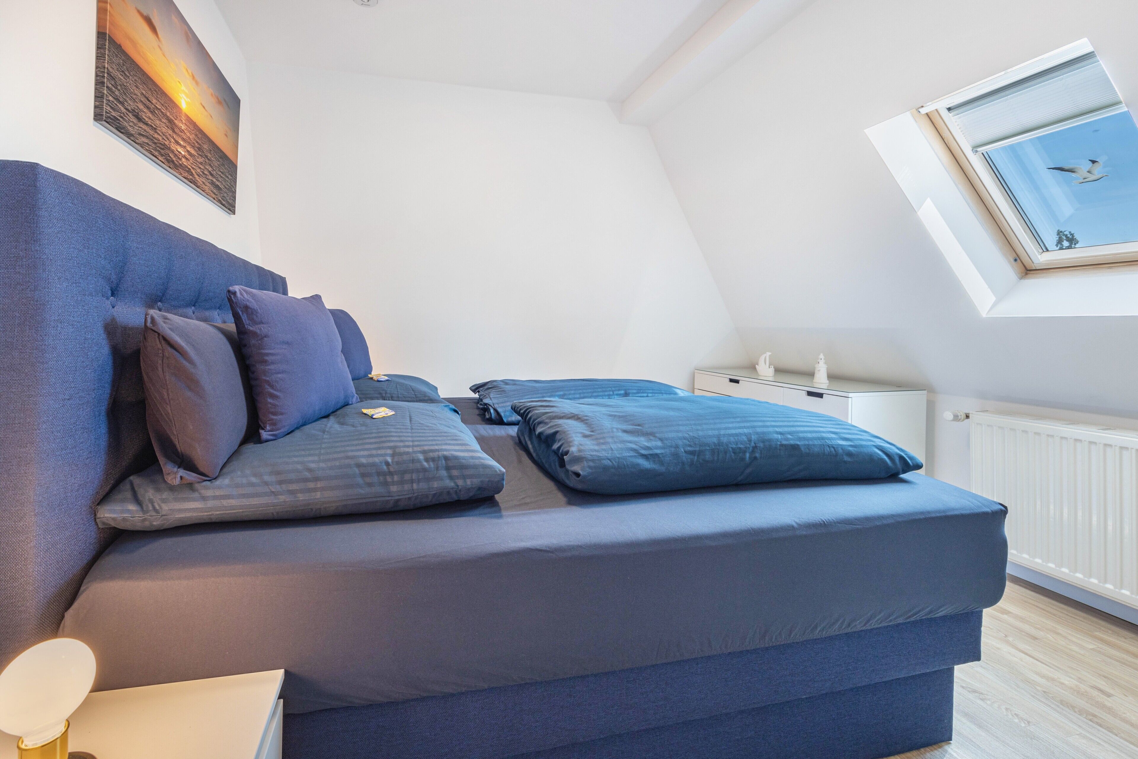 Een slaapkamer met een bed, kussens, een nachtkastje, een raam en een foto aan de muur.