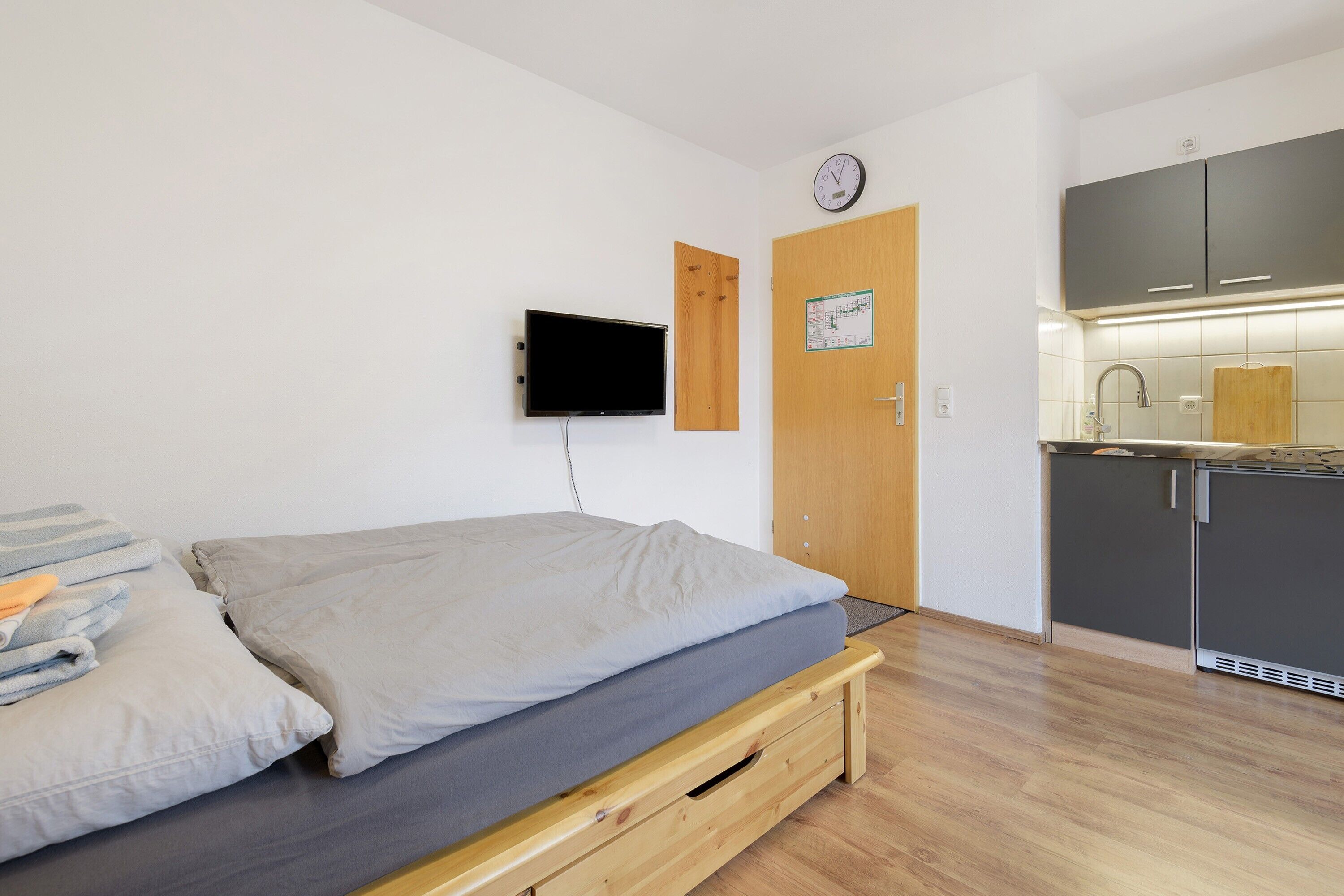 Een kleine kamer met een bed, een aan de muur bevestigde televisie, een houten deur en een kitchenette met kastjes en een spoelbak.