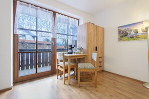 Ein Zimmer mit einem Holztisch, zwei Stühlen, einem Schrank und einem großen Fenster mit Blick auf Gebäude.