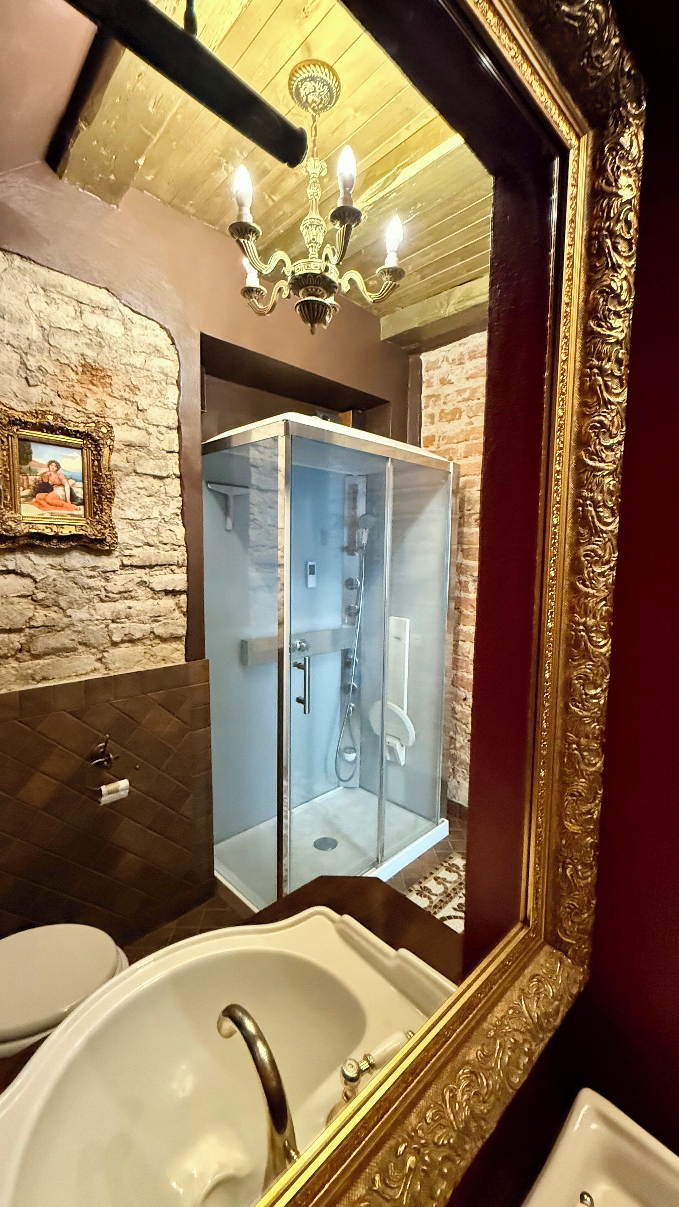 A bathroom with a glass-enclosed shower, a white sink, a mirror with an ornate frame, and a chandelier.