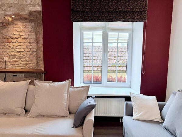 A cozy room with a stone wall, a window with a view of a garden, and a sofa with multiple cushions. - Pilies8 Luxury Vilnius Apartment (Vilnius)