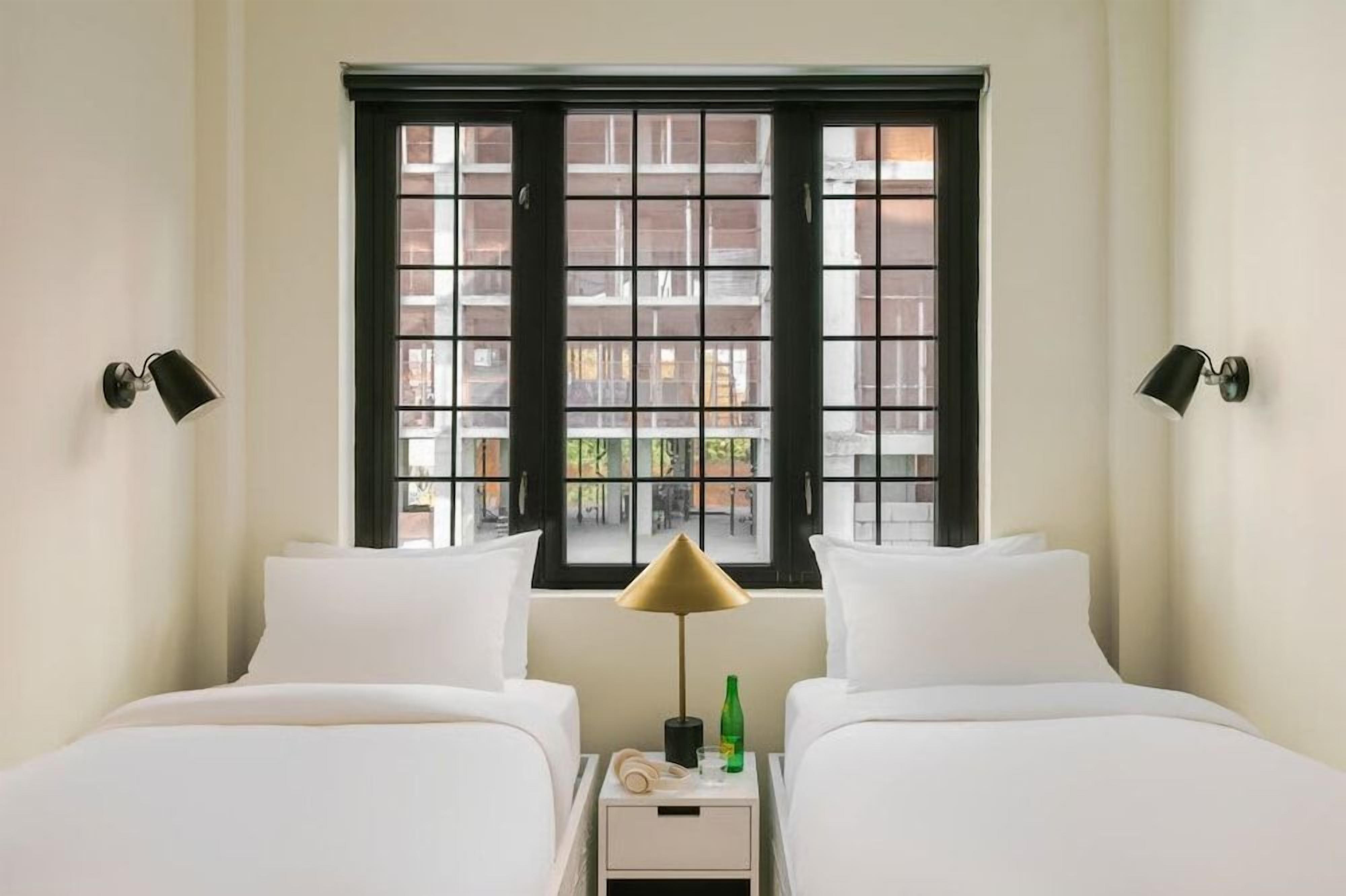 A hotel room with two beds, a window with black frames, and a bedside lamp.
