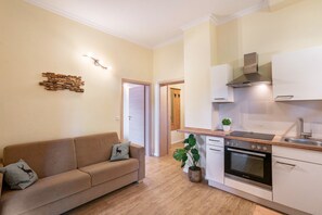 Una cocina moderna con horno empotrado, un lavamanos y una estufa. El suelo es de madera, hay un sofá color beige con cojines decorativos y un aplique de pared.