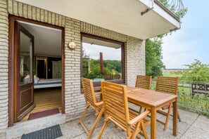 A patio with a table and chairs, a door leading to a room, and a view of greenery.