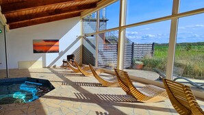 Un área de piscina con tumbonas de madera, suelo de piedra y grandes ventanales con vistas a un edificio y al paisaje.