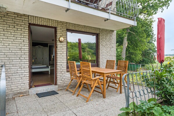 A balcony with a wooden table and chairs, a view of greenery, and a brick wall.