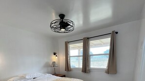 A bedroom with a bed, wooden flooring, a ceiling fan, a window with curtains, and a nightstand with a lamp. - Updated 1BR w/ Kitchen, Fast WiFi + Highway Access Near Carrizo Plain (Cuyama)