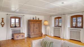 Una habitación con piso de madera, paredes blancas, un sofá blanco con almohadas verdes, un mueble de madera y dos ventanas con cortinas.