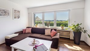A modern living room with a brown sofa, white coffee table, and large windows offering a view of greenery. - Panoramanest Sauerland with Sun Terrace, Near Sorpesee Lake and Hiking Trails (Balve)