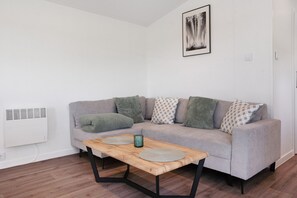 Uma sala de estar com um sofá cinzento, almofadas estampadas, uma mesa de centro de madeira e um quadro na parede.