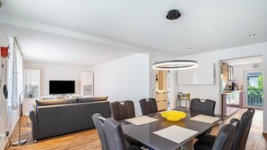 A modern living room with a dining area, a sofa, a TV, and a kitchen in the background.