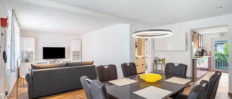 A modern living room with a dining area, a sofa, a TV, and a kitchen in the background.