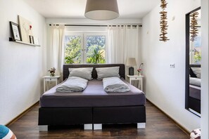 A modern bedroom with a large bed, a window with curtains, a bedside table with a lamp, and a wall-mounted shelf with pictures. - Apartment 'Albi' with Mountain View, Shared Terrace and Wi-Fi (Waldkirch)