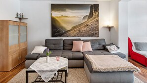 Una sala de estar moderna con un sofá gris, un mueble de madera, una mesa con mantel de encaje y un cuadro de un paisaje montañoso.