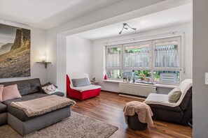 Un salon moderne avec un canapé gris, un fauteuil rouge et un pouf noir. Une grande fenêtre, ornée de rideaux blancs, offre une vue sur la verdure environnante.