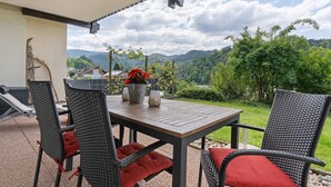 Une terrasse avec des chaises et une table en osier, un pot de fleurs et une vue sur un paysage verdoyant avec des montagnes.