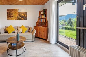 Een woonkamer met een bank, een salontafel, een boekenkast en een groot raam met uitzicht op groen en bergen.