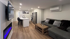 A modern living room with a grey sofa, a wooden coffee table, and a kitchen area with a fridge and a TV. - Cozy 2 bedrooms Suite Near Calgary Airport ✈️ | Air conditioning Sleeps 6 (Calgary)